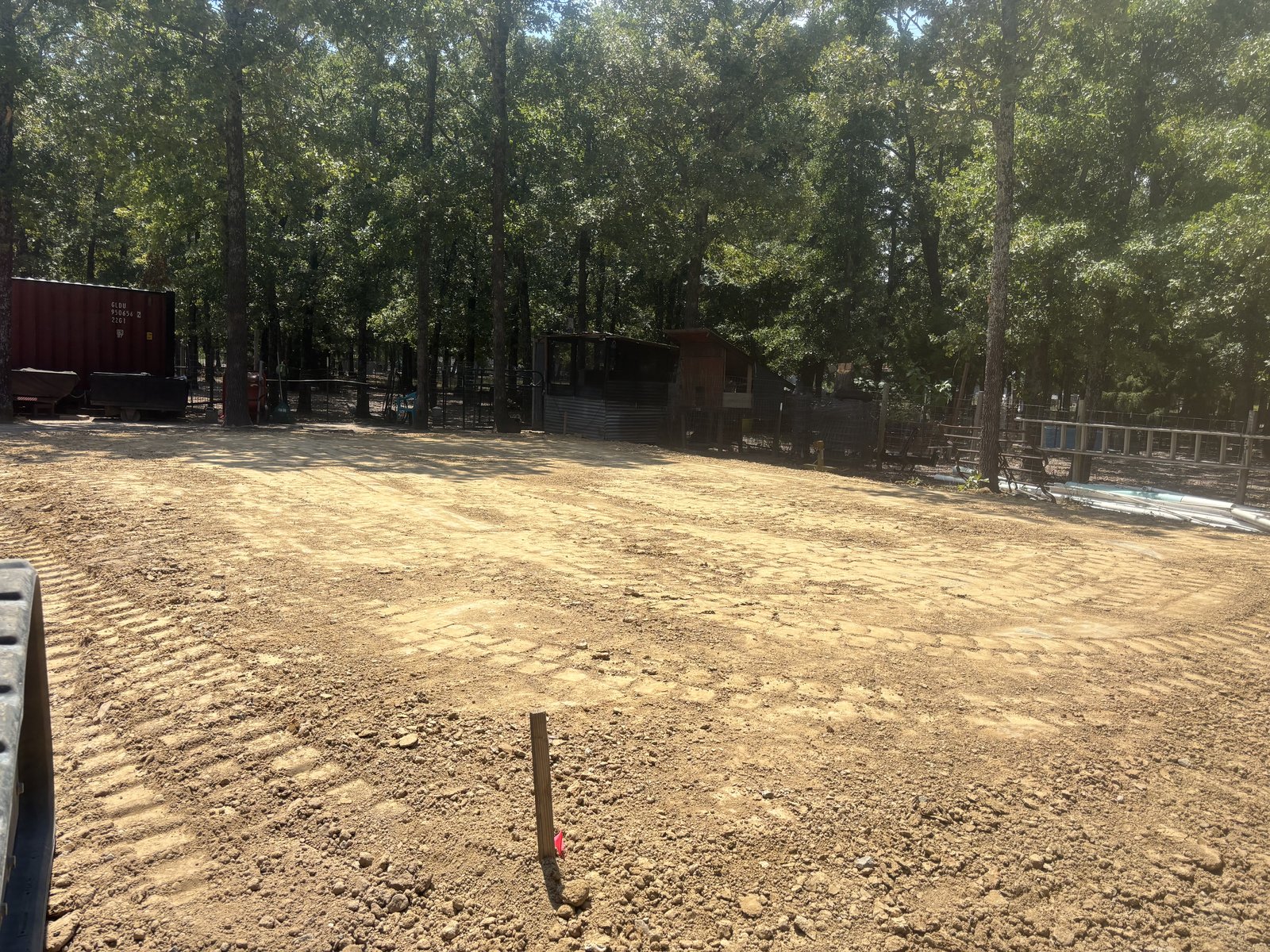 Freshly graded lot with equipment tracks, survey stake, and a shipping container